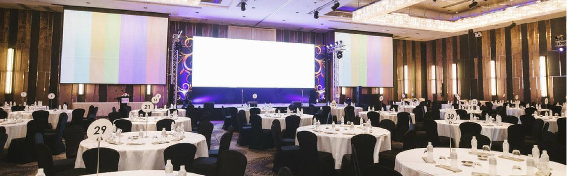 3 large screens with a projector for an event with round tables in the foreground, chairs draped in black.